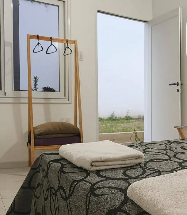 Habitación luminosa con perchero de madera, toallas sobre la cama y puerta que da al jardín